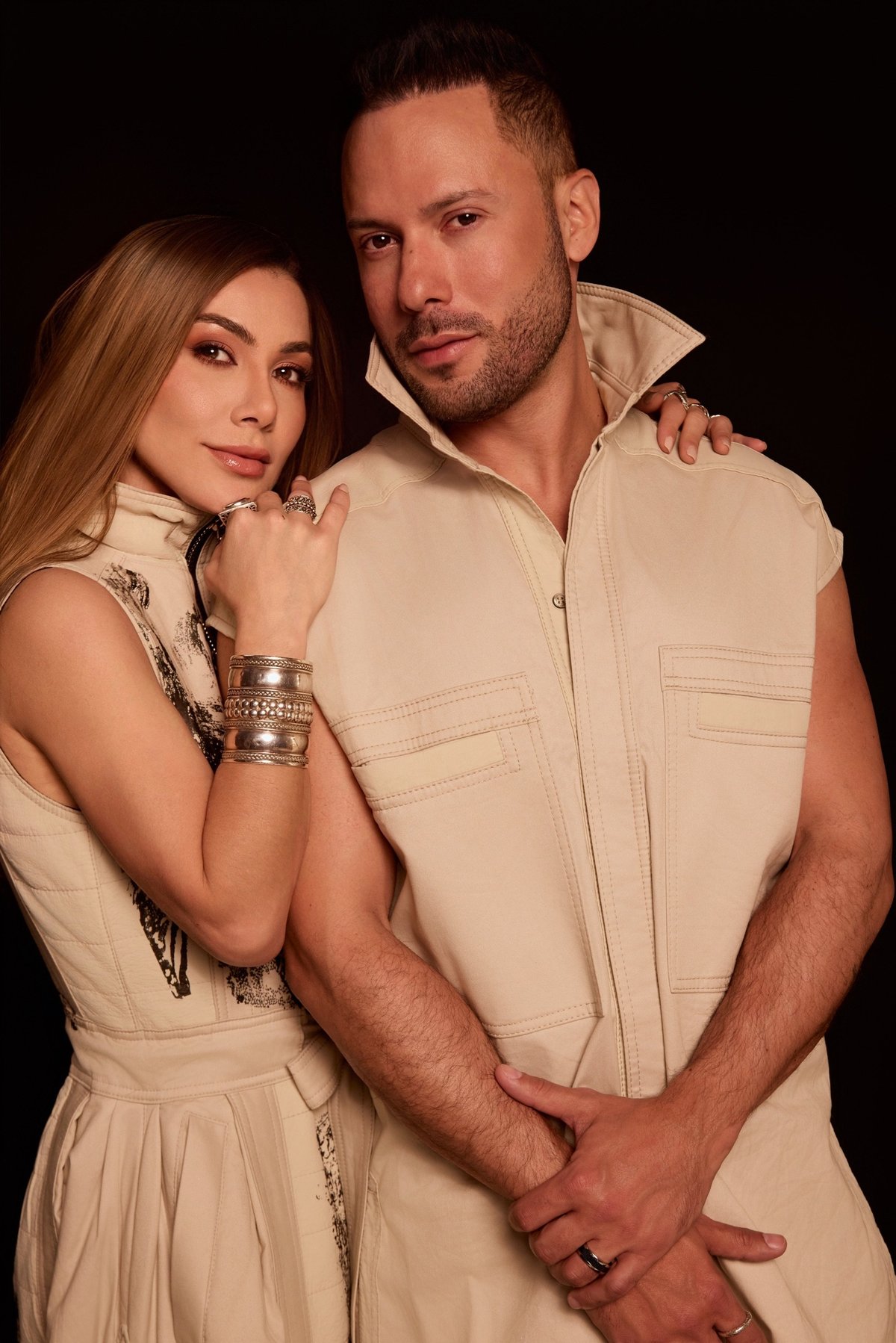 Un hombre y una mujer posando juntos sobre fondo oscuro, ambos con ropa en tonos beige y accesorios metálicos